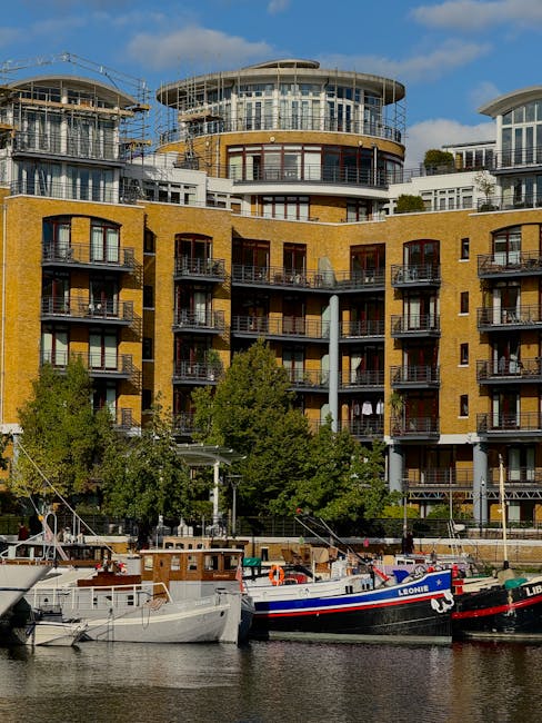 Image depicting a modern residential building along a waterfront, featuring multiple floors with balconies, large glass windows, and a yellow brick exterior. The upper floors include circular and roun