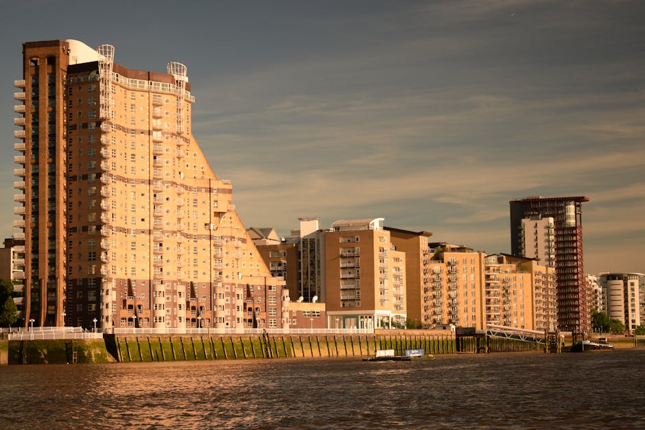 Image depicting a modern residential building along a waterfront, featuring multiple floors with balconies, large glass windows, and a yellow brick exterior. The upper floors include circular and roun