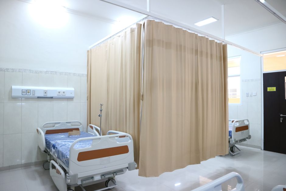A hospital bed with a metal frame and adjustable headrest, situated near a large window with sheer white curtains allowing diffuse natural light into the room. The bed has striped bedding in shades of